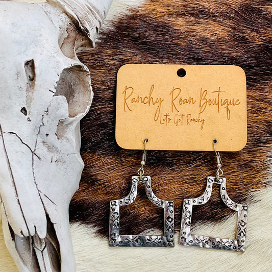 Western Cattle Tag Cutout Dangle Earrings displayed on a ranchy background with cowhide and skull decor.