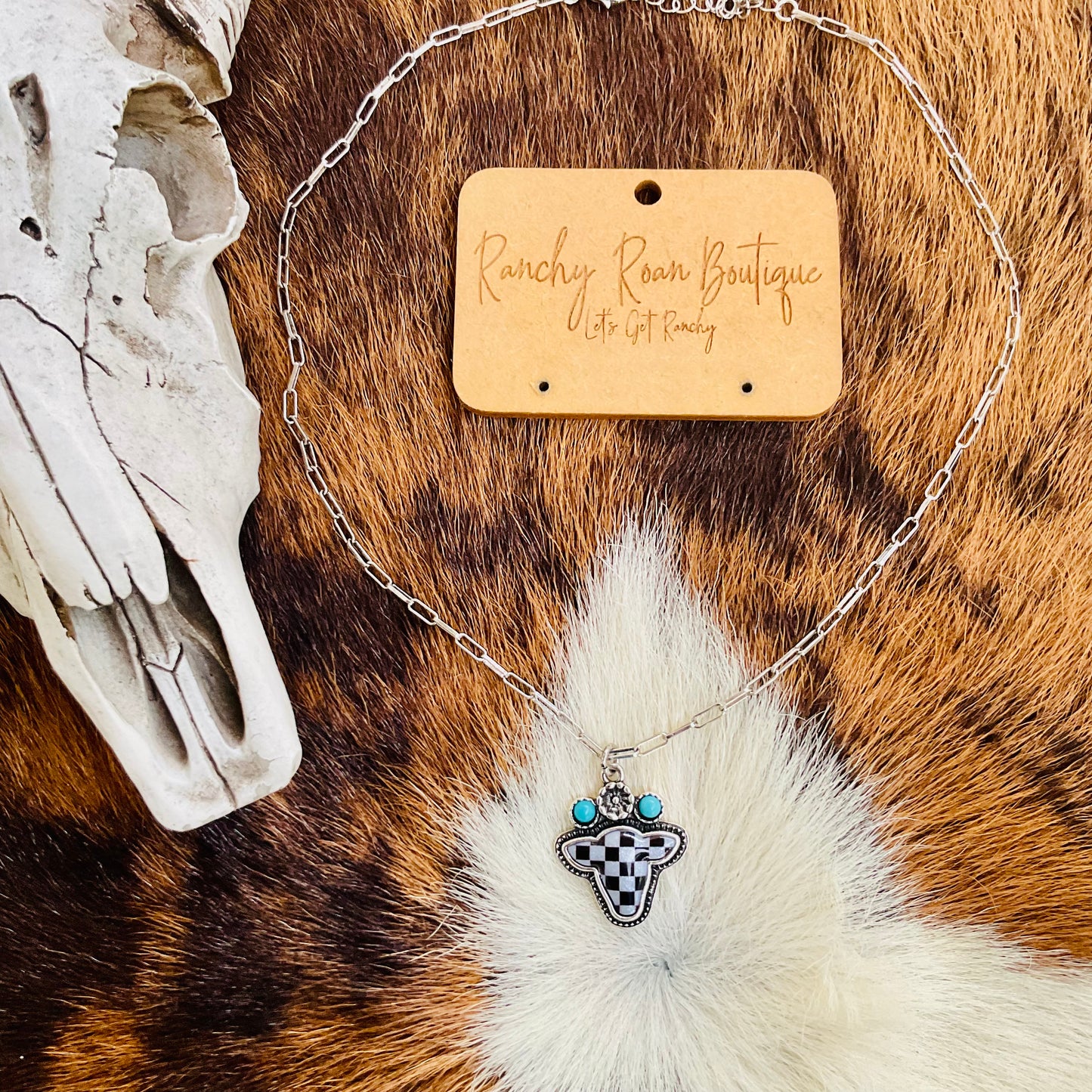Western Checker Cow Head Necklace displayed on a fur backdrop, featuring a silver-tone pendant with a checker pattern and turquoise details.