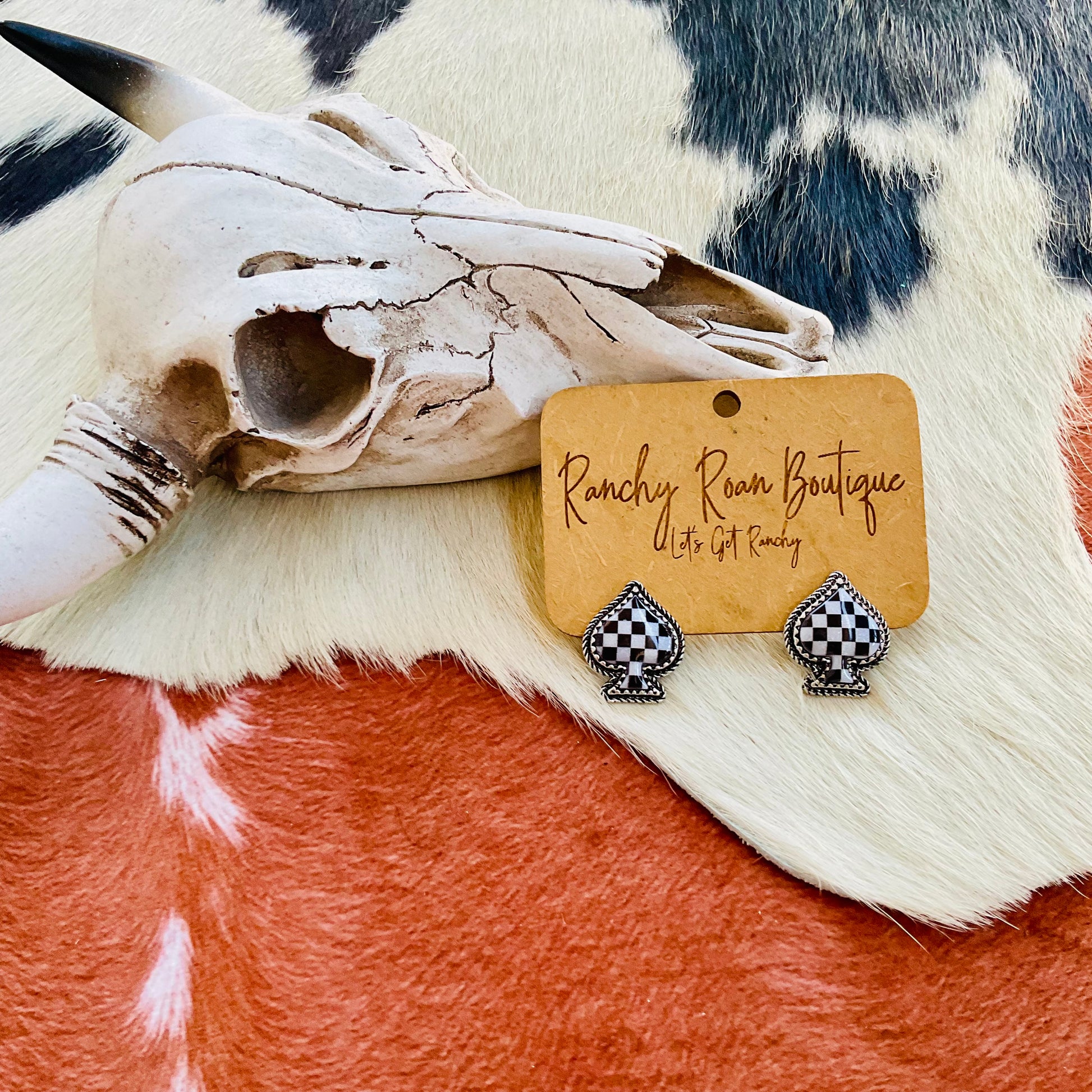 Checker-pattern spade-shaped stud earrings displayed on a Ranchy Roan Boutique tag, set against a Western-style cowhide background with a decorative skull.