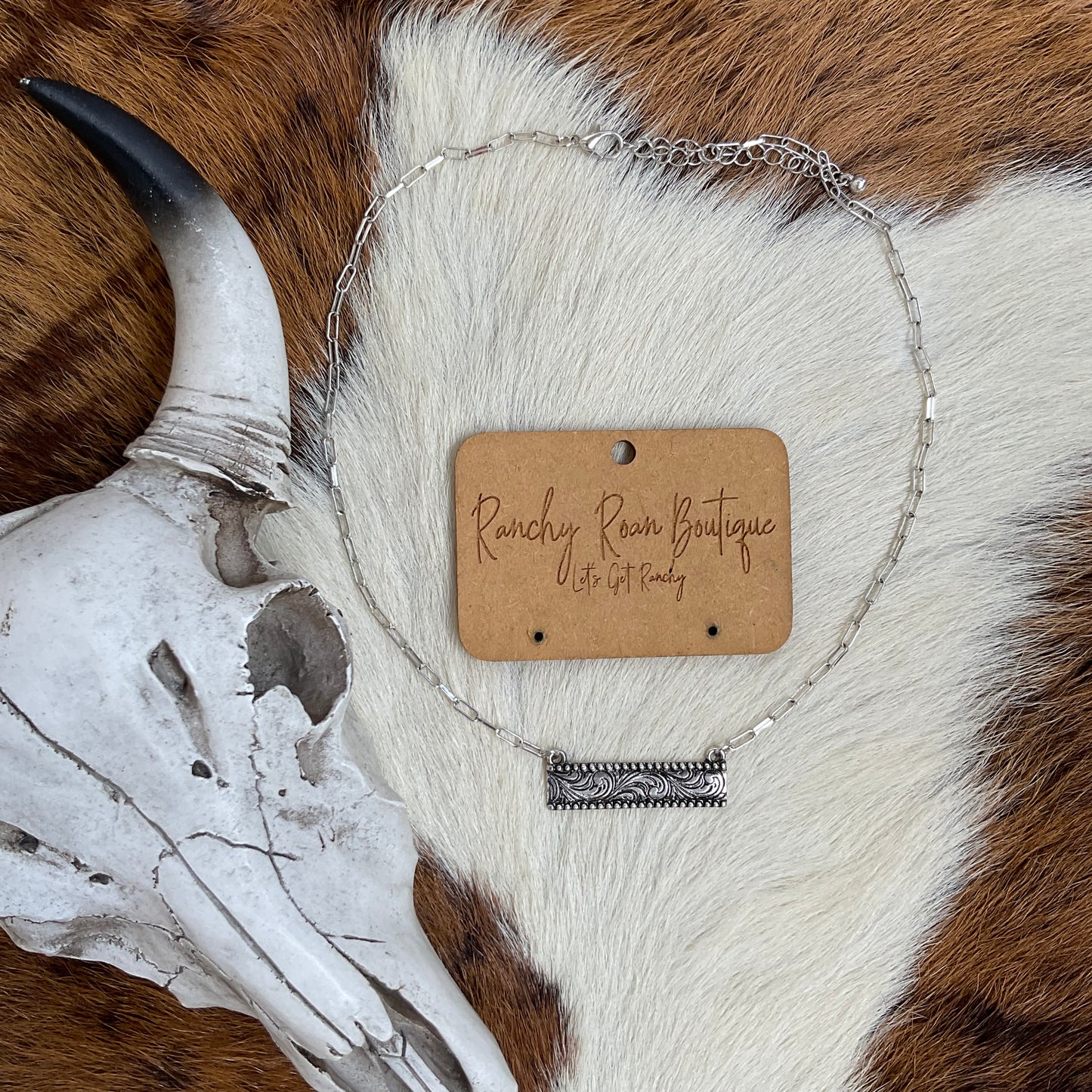  Western Dainty Bar Necklace displayed on a soft cowhide backdrop, showcasing its intricate engraved bar pendant and adjustable chain.
