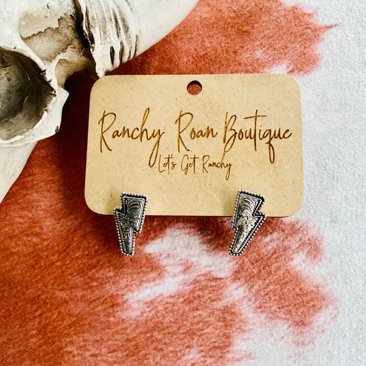 Western Dainty Thunder stud earrings in a lightning bolt shape with burnished silver finish, displayed on a Ranchy Roan Boutique card against a rustic background.