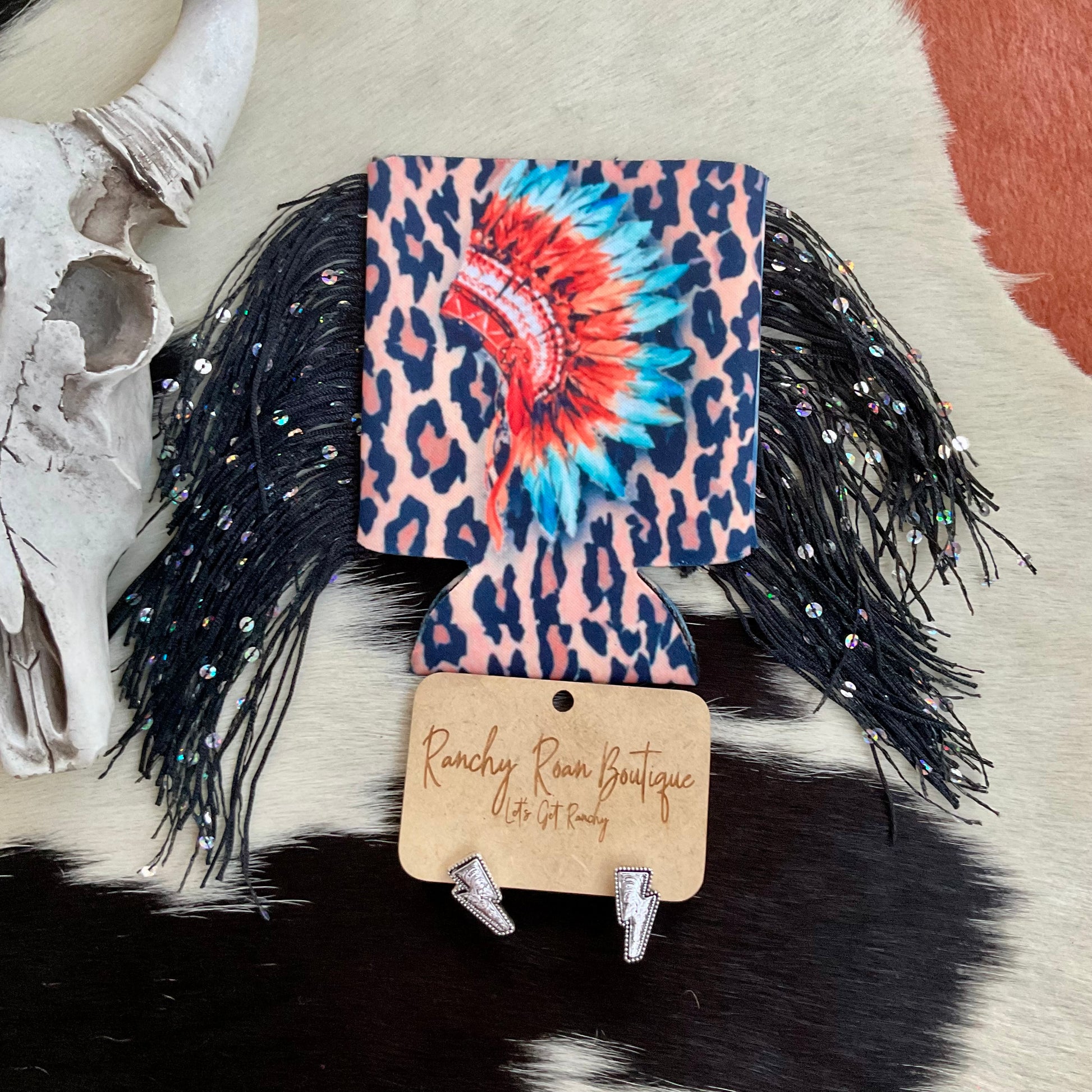 A leopard print koozie with a vibrant Western headdress design, featuring black fringe detailing on a Western backdrop.