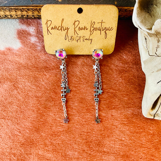 Close-up of Western wild typography stud earrings featuring an AB stone and chain drop with star accents, displayed against a rustic background with cow skull decor.