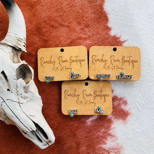 Western Word Stud Earrings featuring the words “Cowgirl,” “Howdy,” and “Yeehaw” with turquoise accents, displayed with a decorative skull on a cowhide rug.