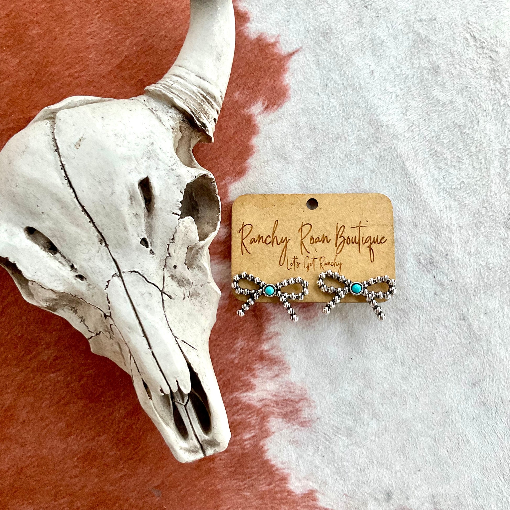 Burnished silver ribbon stud earrings with a turquoise center, displayed on a Ranchy Roan Boutique card against a cowhide background.