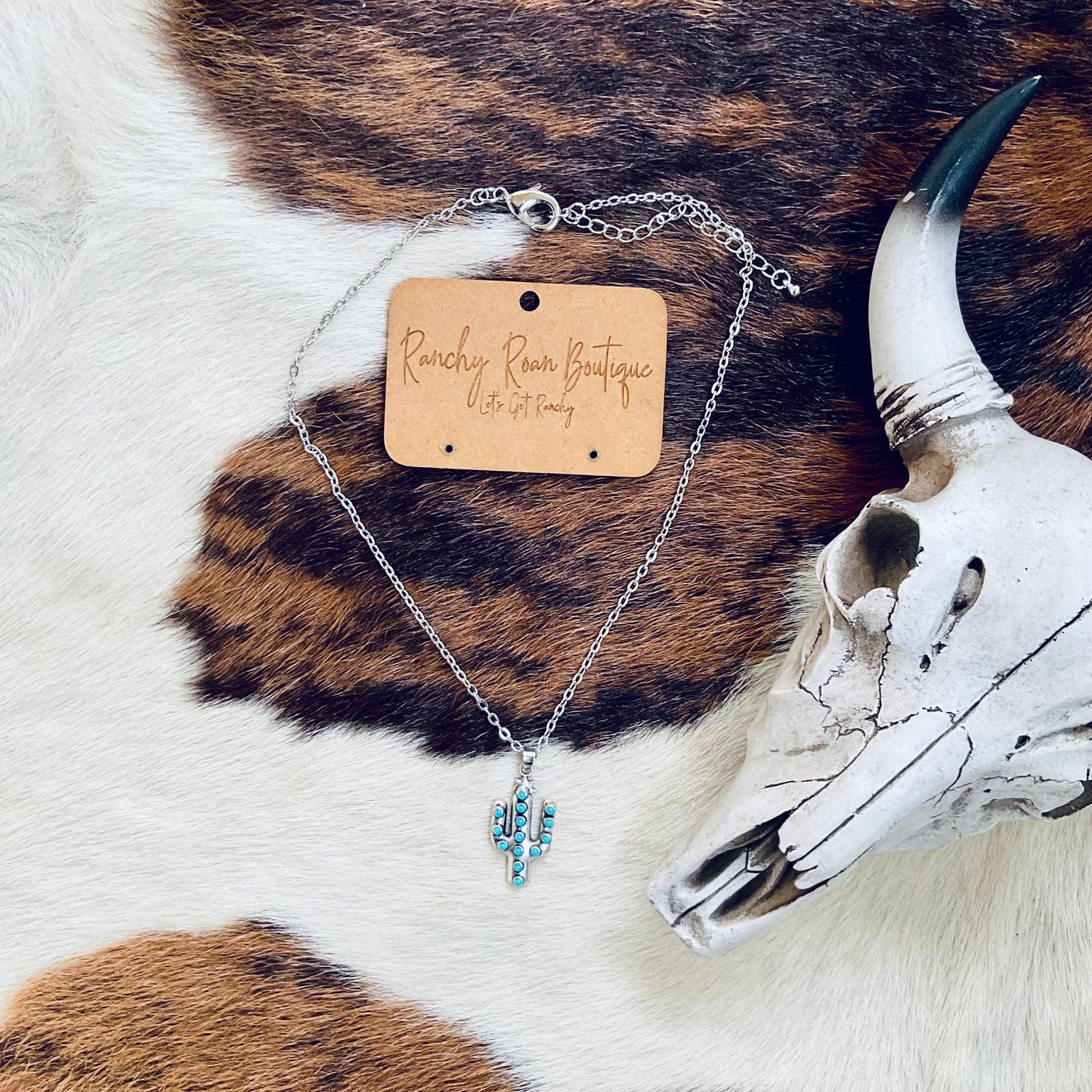 Close-up of a turquoise cactus pendant necklace styled on a cowhide background next to a Western-themed decor skull.