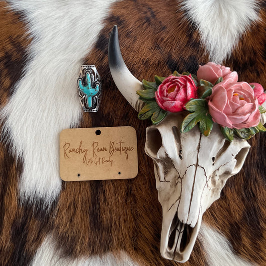 Front view of a turquoise cactus Western ring in a silver-tone setting, displayed with a decorative skull on a cowhide background.